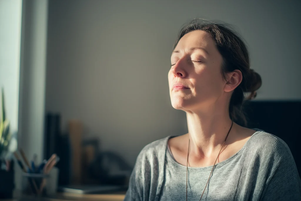 Adulte assis à un bureau à domicile, respirant calmement, les yeux fermés, dans une ambiance lumineuse et apaisante.