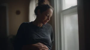 Femme enceinte au tout début de grossesse debout près d’une fenêtre posant les deux mains sur son bas-ventre encore discret dans une lumière naturelle douce