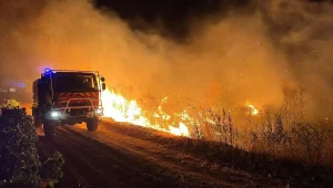 Colline boisée en feu à l'horizon sous une fumée orangée, une personne ferme les volets de sa maison en fin d'après-midi.